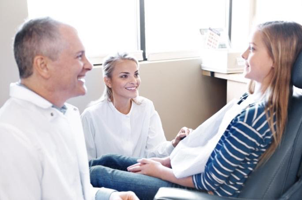 A dentist and his dental assistant make a young female patient laugh in the dental chair.